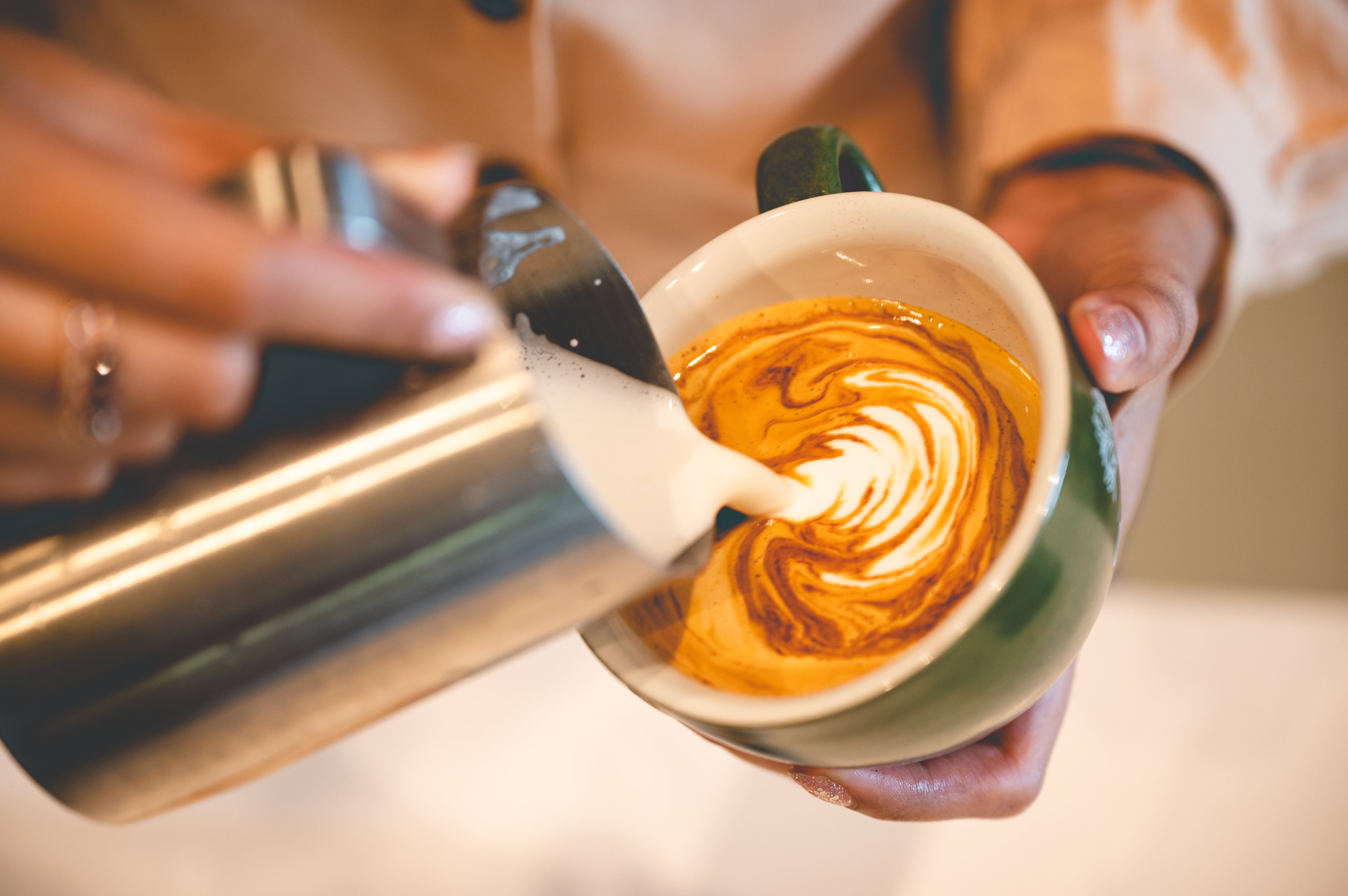 A close-up of a barista pouring steamed milk into a cup of espresso to create latte art, forming a detailed rosette pattern on the coffee surface.
