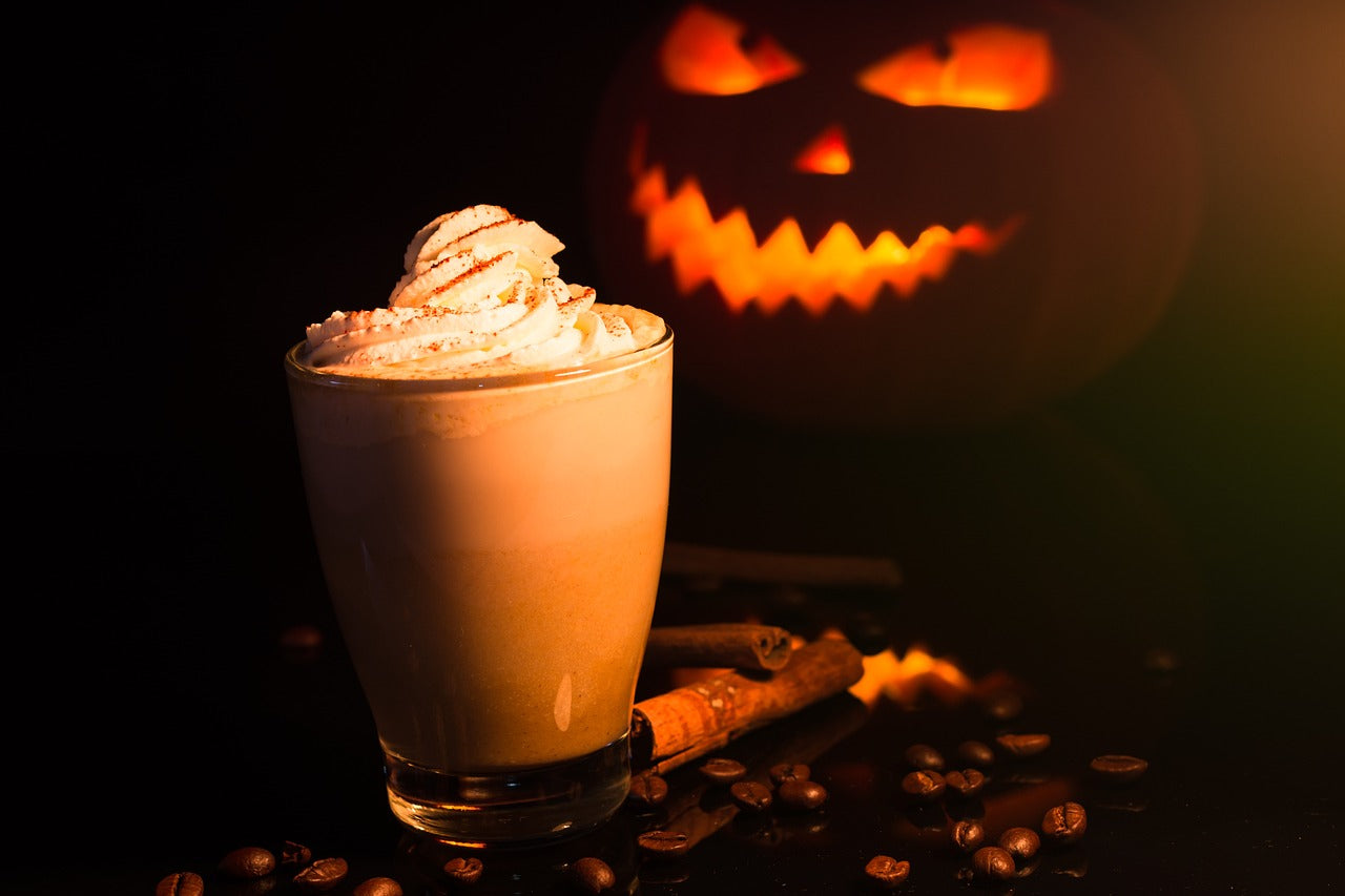 A festive pumpkin spice latte topped with whipped cream, surrounded by cinnamon sticks and coffee beans, with a glowing carved Halloween pumpkin in the background.