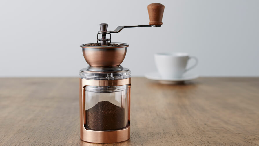 Manual coffee grinder filled with freshly ground coffee on a wooden table, with a white cup and saucer in the background.