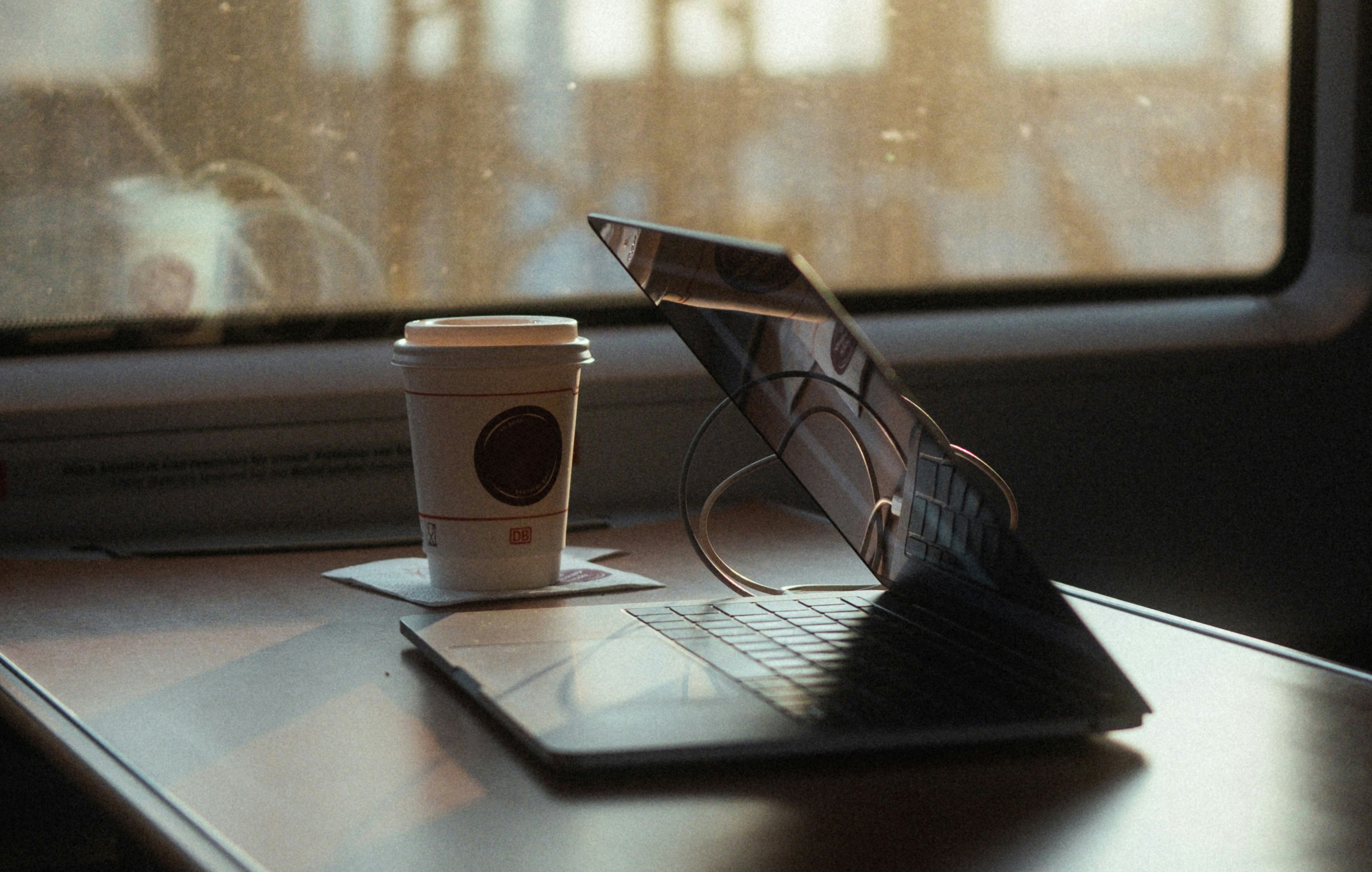 A takeaway coffee cup and open laptop resting on a train table, with soft natural light streaming through the window, capturing a peaceful travel or remote work moment.